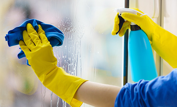 a cleaner wearing yellow gloves spraying and wiping a window with a blue bottle and cloth